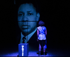 Comédienne faisant face à un grand portrait projeté sur scène, dans une scénographie évoquant la mémoire et l’histoire dans le spectacle « J’ai plusieurs terres »