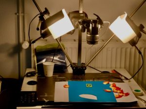 Caméra de tournage stop-motion fixée au-dessus d'une table éclairée avec des décors en carton en forme de flammes.