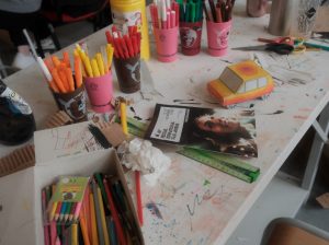 Table d'atelier créatif encombrée de feutres colorés, de ciseaux et d'une petite voiture en carton plié.