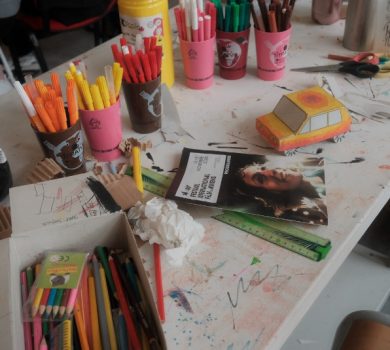 Table d'atelier créatif encombrée de feutres colorés, de ciseaux et d'une petite voiture en carton plié.